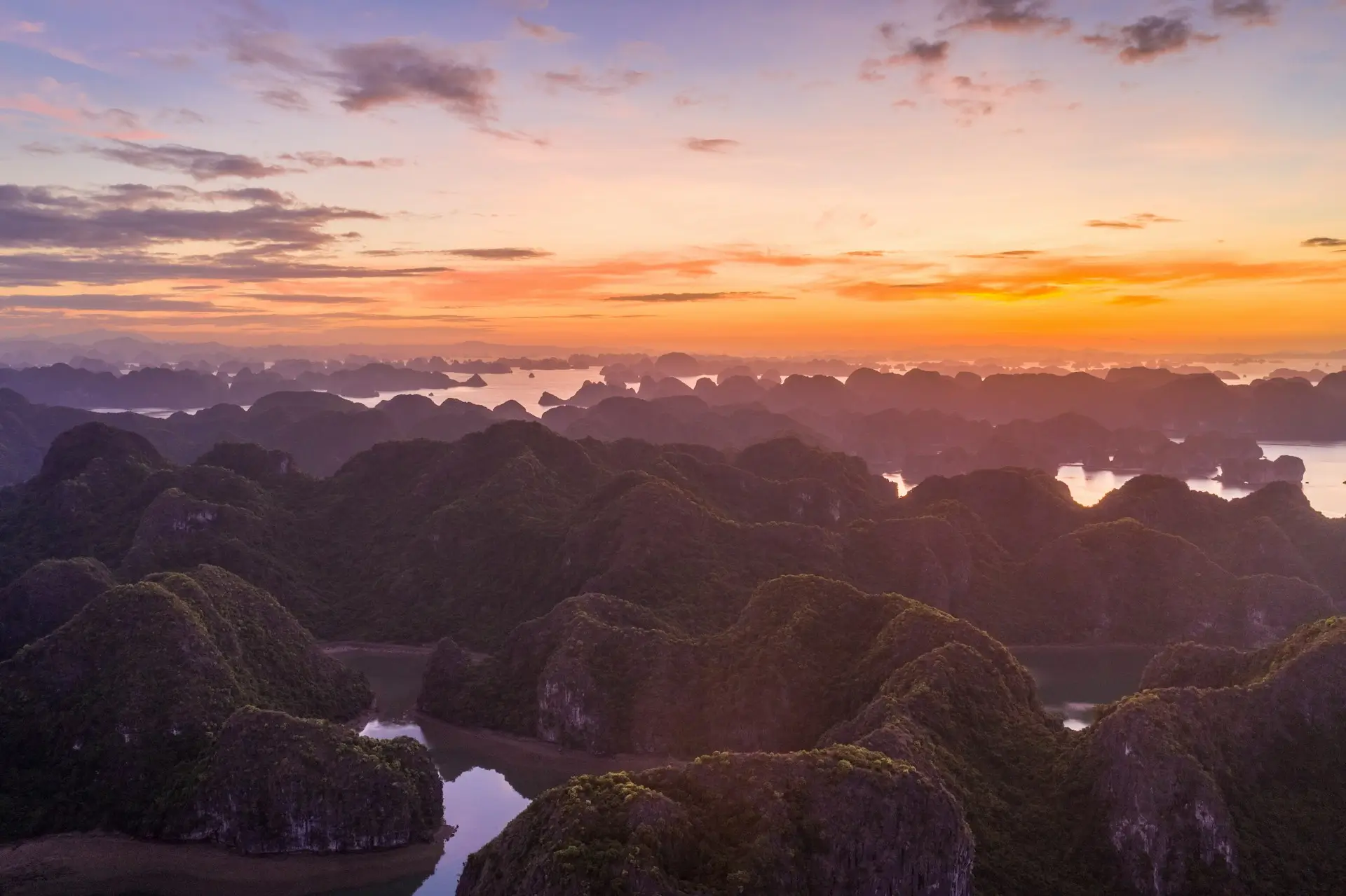 Vista mozzafiato del tramonto con sfumature arancioni e violacee che illuminano il cielo sopra la Baia di Ha Long in Vietnam, riconosciuta dall'UNESCO per il suo straordinario patrimonio naturale, mostrando isolotti calcarei e acque tranquille, ideale per promuovere il turismo sostenibile e la valorizzazione culturale nella regione di Quang Ninh.