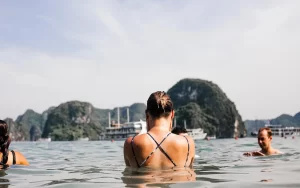 Una viaggiatrice solitaria nuota nella Baia di Ha Long in Vietnam, con le famose formazioni calcaree sullo sfondo, ideale per il turismo eco-sostenibile e le avventure individuali.