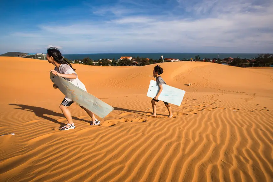 Due bambini che fanno sandboarding sulle dune di sabbia di Mui Ne, in Vietnam, portando le loro tavole mentre camminano sulle dune.