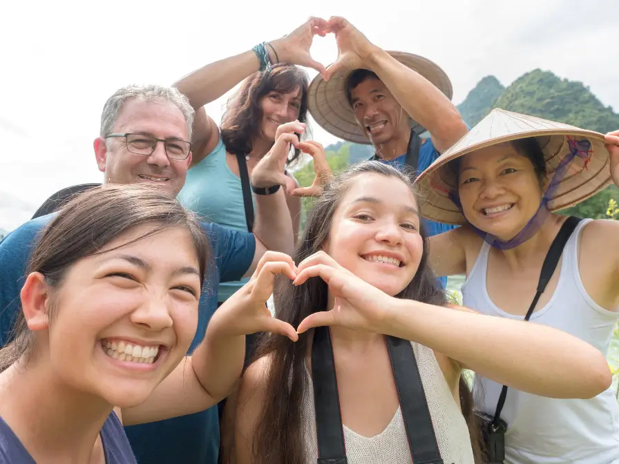 Un gruppo di amici sorridenti che fanno il segno del cuore con le mani durante un tour in Vietnam, indossando cappelli tradizionali.