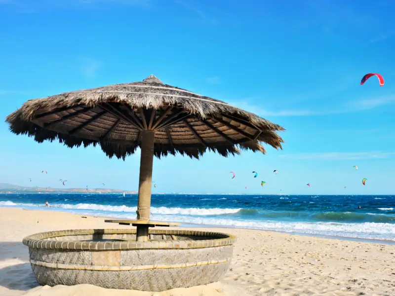 Spiaggia di Mui Ne in Vietnam con ombrelloni di paglia e kite surfer che volano sopra le onde, ideale per sport acquatici e relax